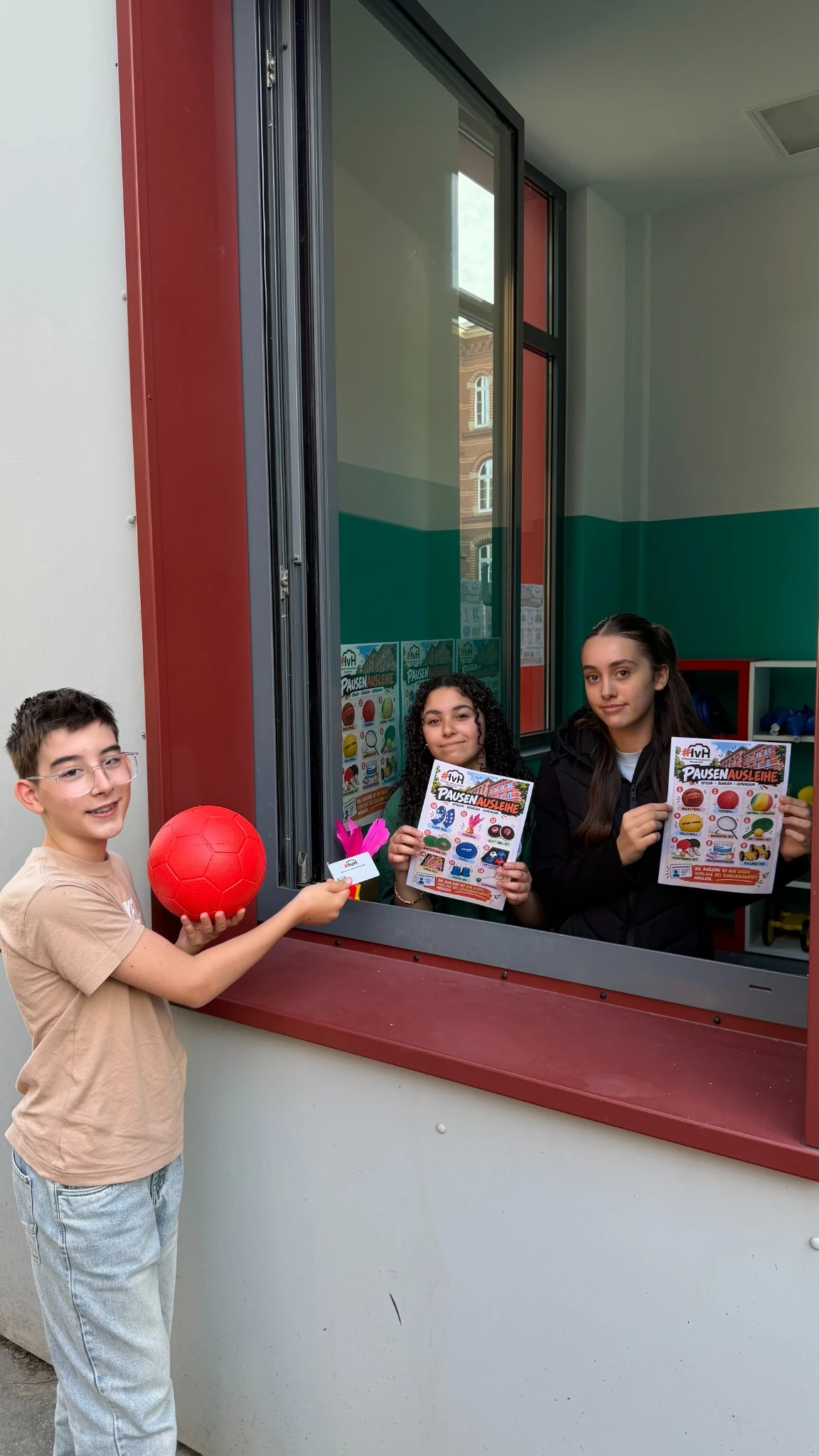 Ein Schüler zeigt seinen Ausweis am Ausgabefenster. Zwei Schülerinnen halten die Übersichten der Spielgeräte bereit.